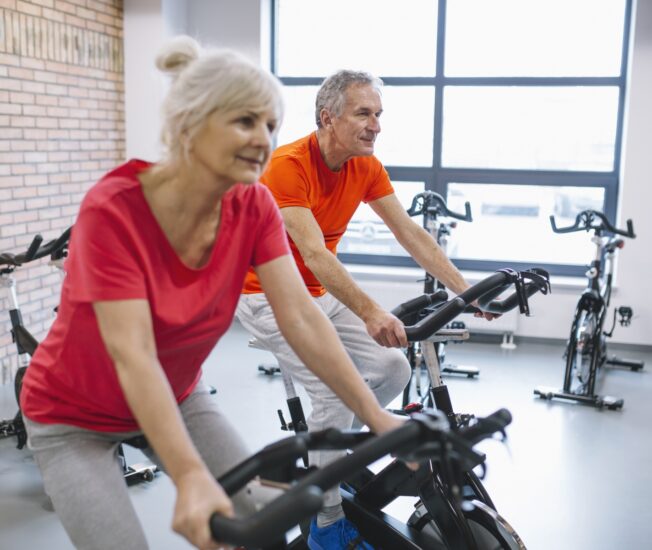 En äldre kvinna och en äldre man sitter på varsin motionscykel i ett gym.