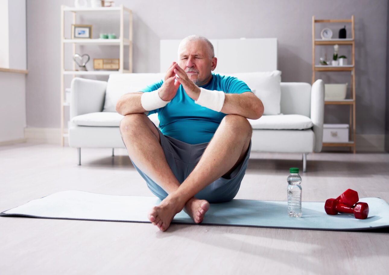 Äldre man sitter med benen i kors på en yogamatta i ett vitt vardagsrum.
