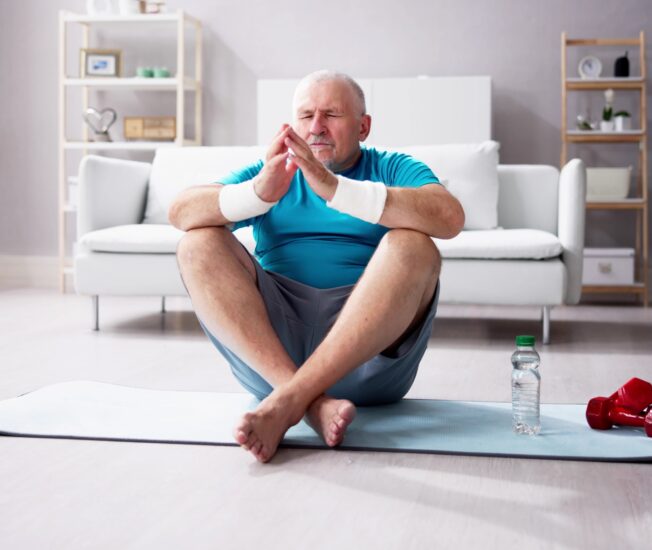 Äldre man sitter med benen i kors på en yogamatta i ett vitt vardagsrum.