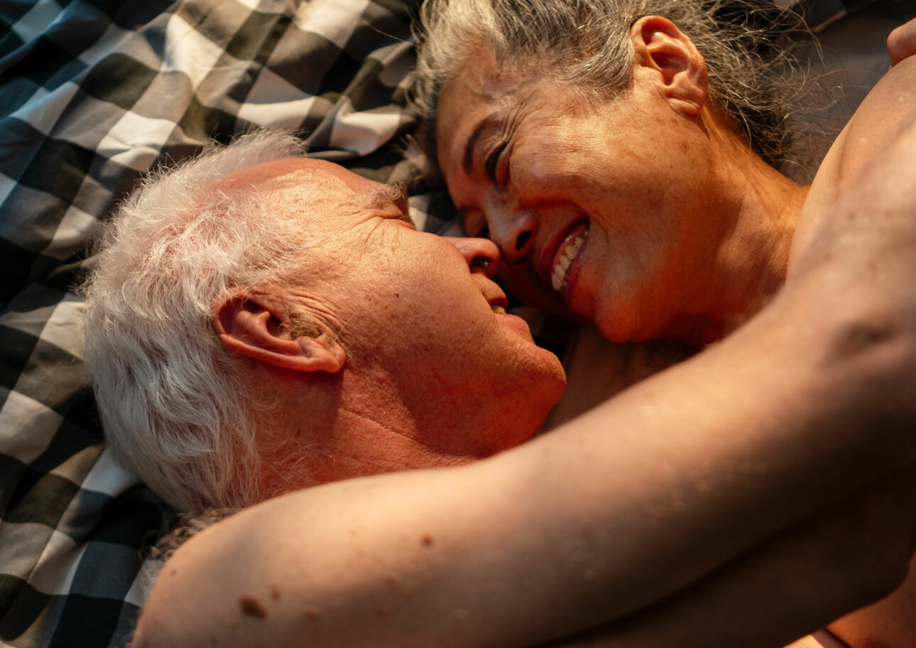 Äldre naket par omfamnar varandra i en säng och ler lyckligt mot varandra.