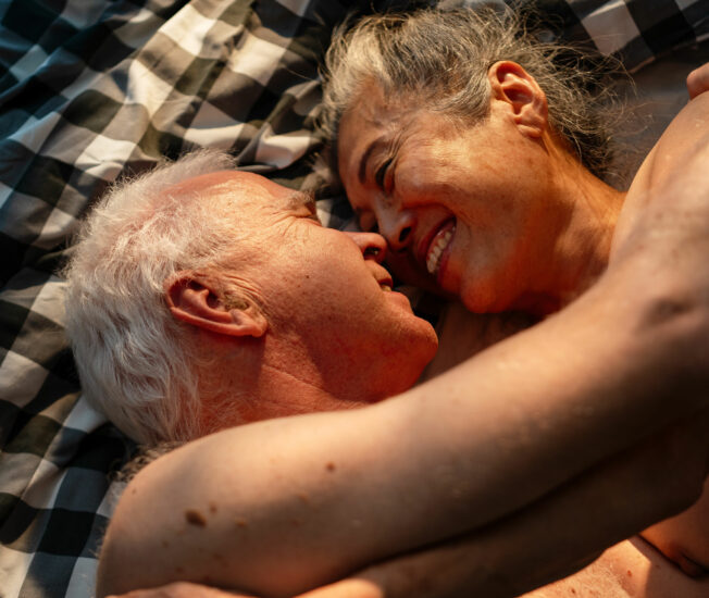 Äldre naket par omfamnar varandra i en säng och ler lyckligt mot varandra.