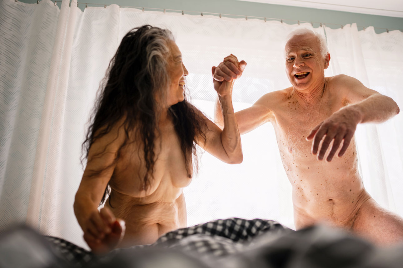 Kvinna och man, båda äldre och nakna, i ett sovrum håller varandras hand och tar ett skutt ner i en säng.