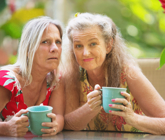 Två äldre kvinnor sitter tätt tillsammans med muggar i händerna blickar oroligt och misstänksamt mot kameran