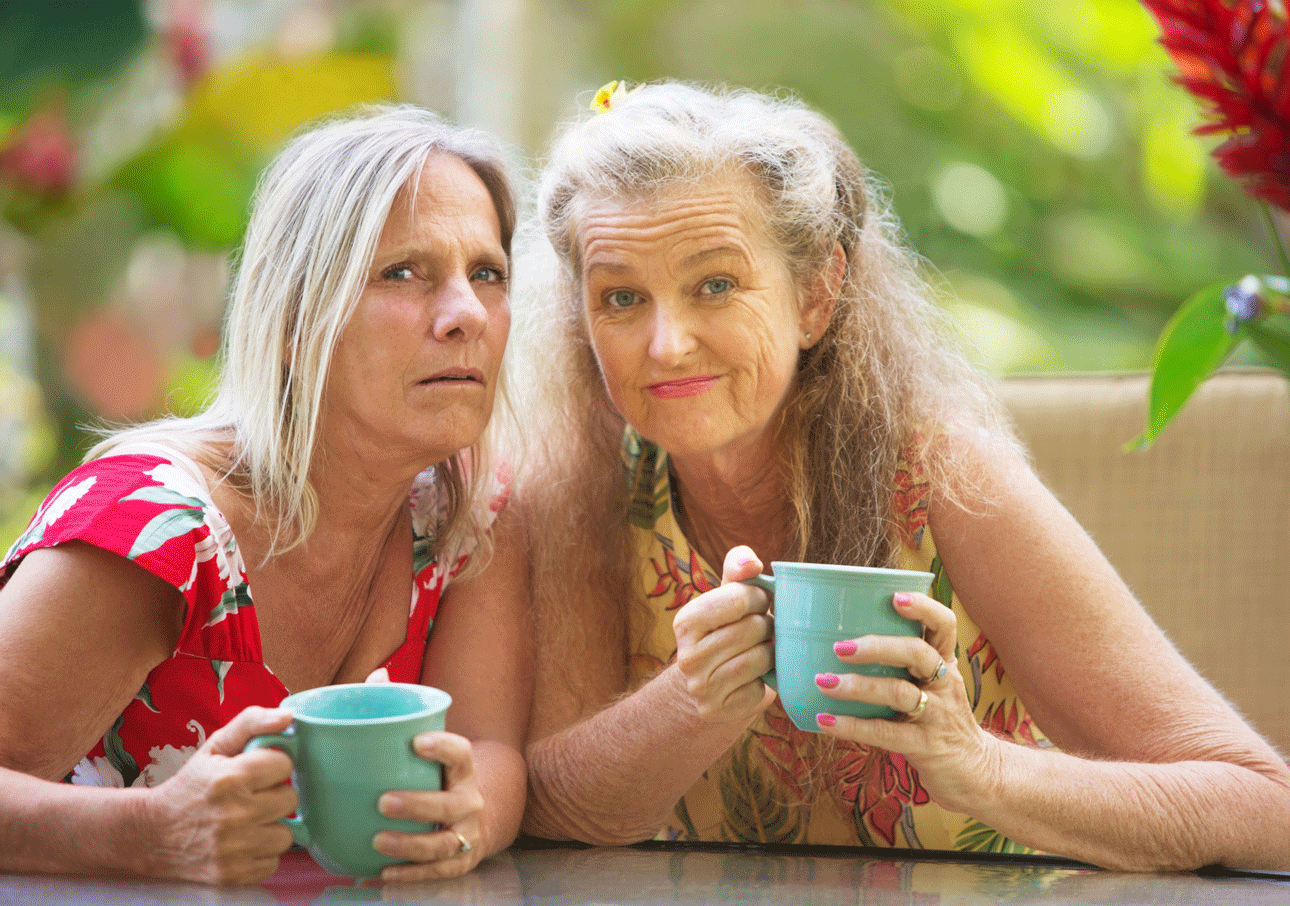 Två äldre kvinnor sitter tätt tillsammans med muggar i händerna blickar oroligt och misstänksamt mot kameran