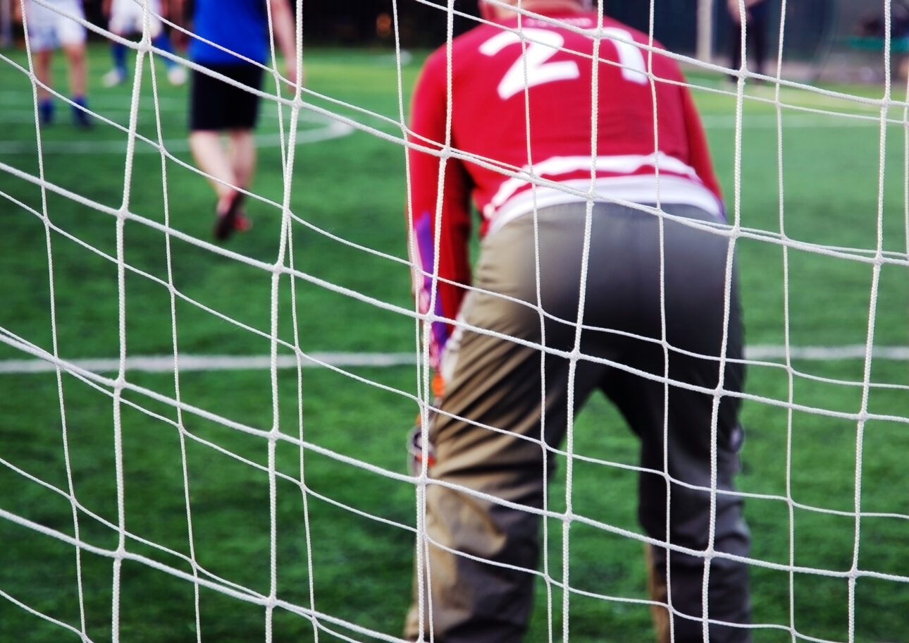 Foto som visar ryggen på en fotbollsmålvakt med fotbollsplanen i bakgrunden.