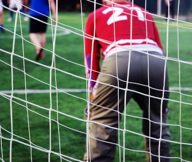 Foto som visar ryggen på en fotbollsmålvakt med fotbollsplanen i bakgrunden.