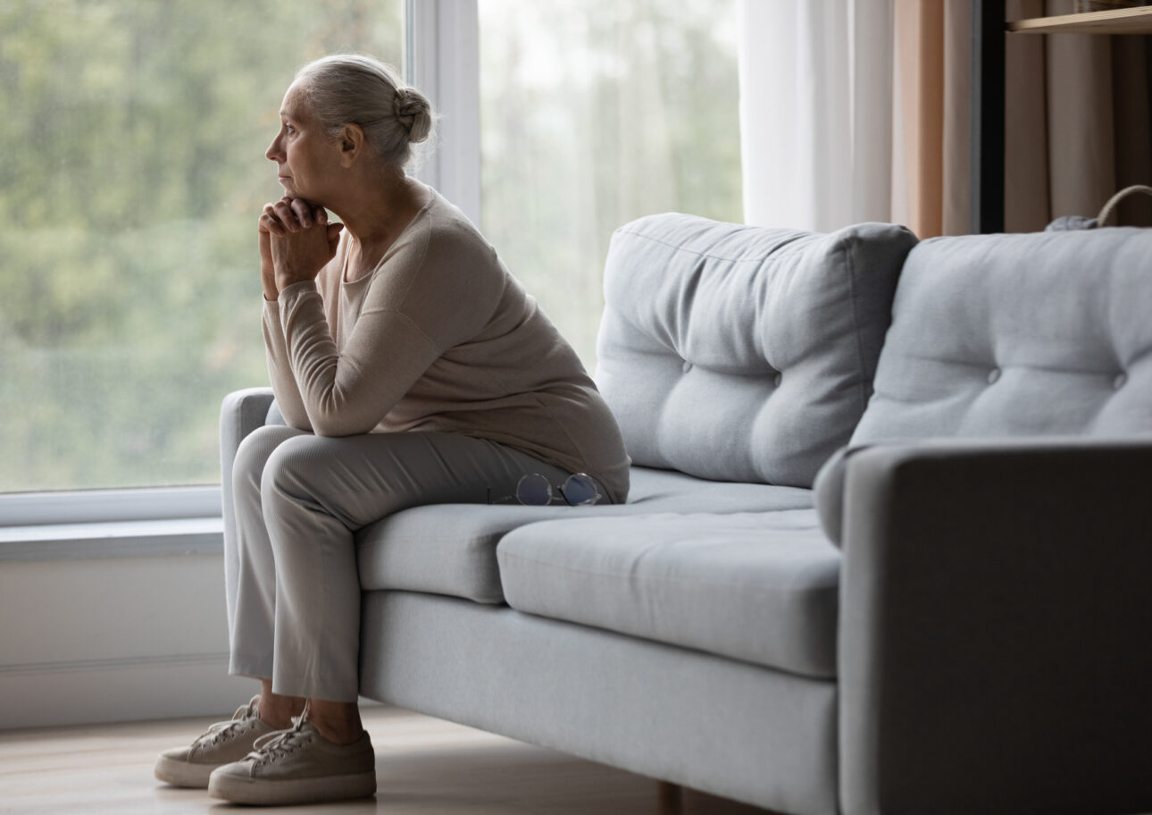 Äldre kvinna sitter i en soffa och tittar ut genom fönstret.