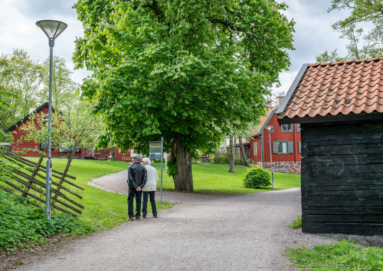 Två äldre kvinnor går tillsammans på en grusgångväg vid flera faluröda hus, Färgaregården i Norrköping.