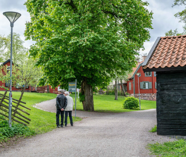 Två äldre kvinnor går tillsammans på en grusgångväg vid flera faluröda hus, Färgaregården i Norrköping.