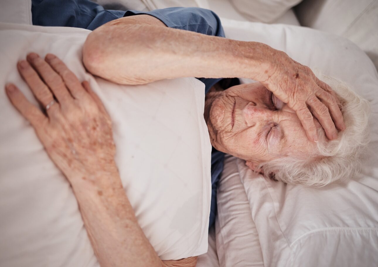 Äldre kvinna ligger i sängen och håller sig om huvudet