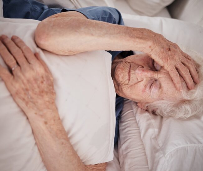 Äldre kvinna ligger i sängen och håller sig om huvudet